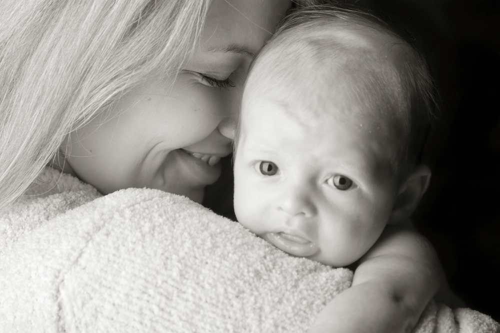 Black and white closeup image of a mother smiling while holding her baby over shoulder